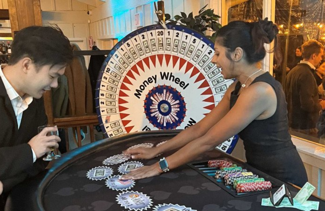 A money wheel spins in the background while a dealer lays funny money down on a table for  a player who observes smiling with a drink in his hand.