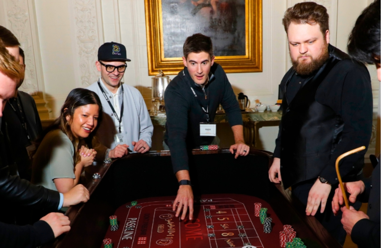 a man throws dice on a high end craps table while a group of participants watch in excitement at a casino night fundraiser