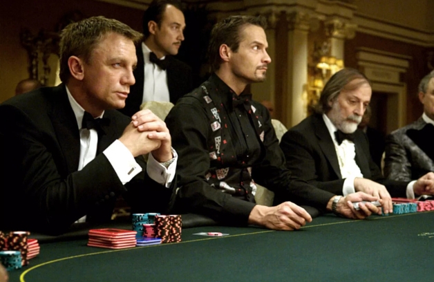 a movie scene from james bond casino royale depicts men siting around a casino table dressed in bond themed costume outfits