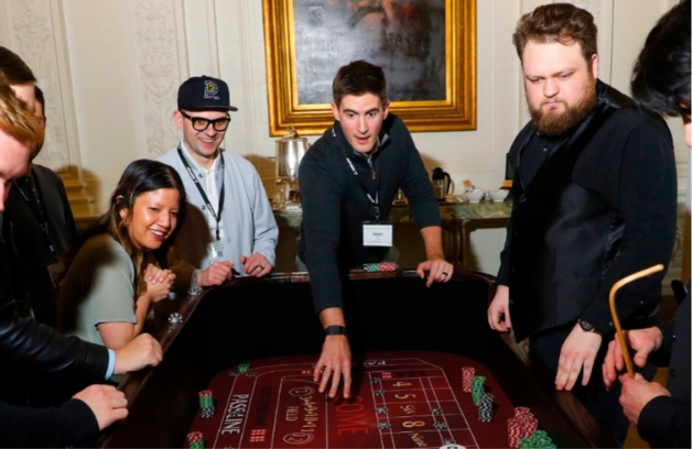 a man throws dice on a high end craps table while a group of participants watch in excitement at a casino night fundraiser