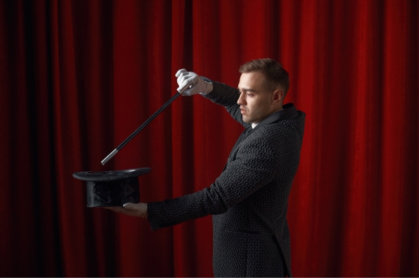magician pointing his magic wand at his hat with gloved hands in front of a red backdrop